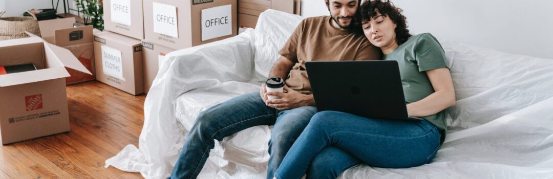 couple sitting on couch using laptop with moving boxes near by